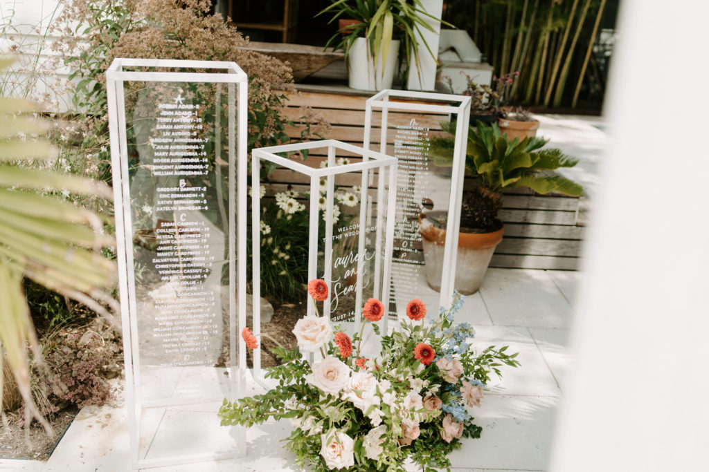 Three clear acrylic & painted wood columns by an NJ wedding calligrapher displaying a seating chart and welcome sign for a wedding with a floral arrangement on the floor in front.