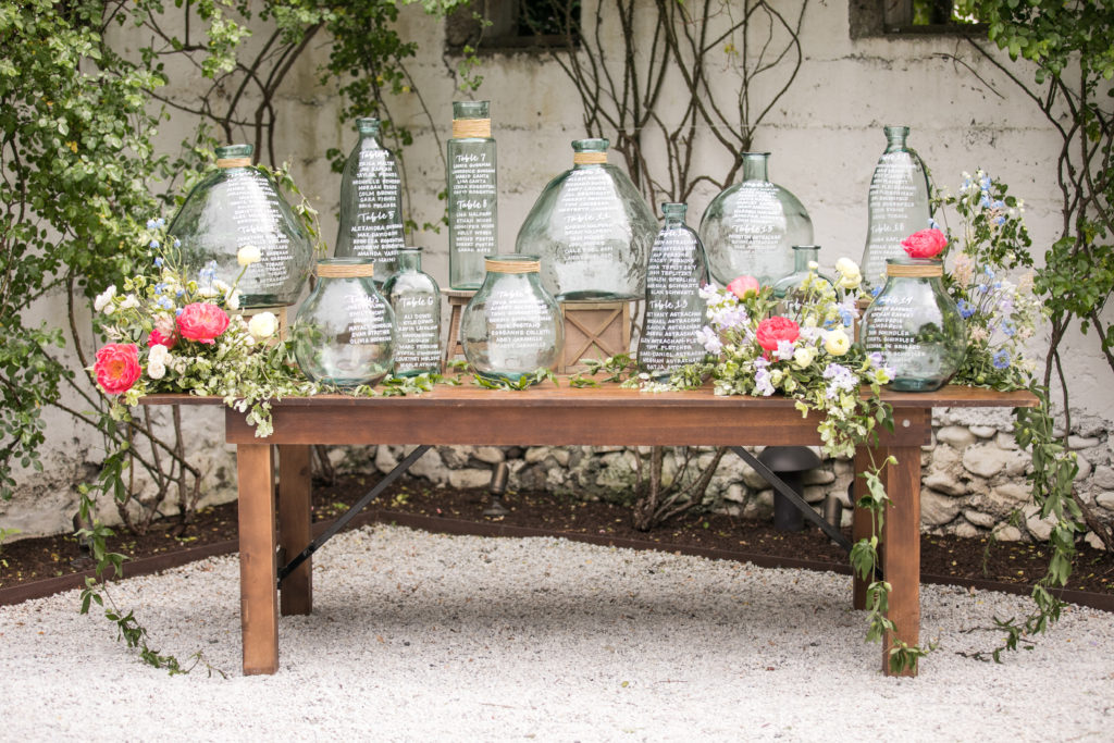 Glass vase seating chart by NJ wedding calligrapher displayed on farmhouse table with greenery and florals.
